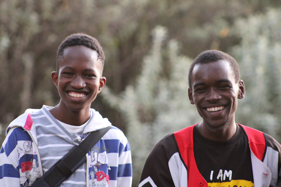 In a serene park setting, two young men are captured in a moment of joy and camaraderie. The man on the left is wearing a blue and white striped shirt with a black backpack slung over his shoulder, while the man on the right sports a red and yellow jacket adorned with a "I AM" design.

Both men have their mouths open wide, as if they are caught mid-laughter or sharing an inside joke. The background of the image reveals a tranquil park scene, with trees standing tall in the distance and bushes dotting the landscape.
