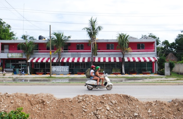 A pink and white two-story hotel with red and white striped awnings is surrounded by palm trees. A scooter with two riders, both wearing helmets, travels on the road in front of the hotel. The ground in the foreground is covered with dirt, suggesting construction work.