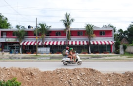 A pink and white two-story hotel with red and white striped awnings is surrounded by palm trees. A scooter with two riders, both wearing helmets, travels on the road in front of the hotel. The ground in the foreground is covered with dirt, suggesting construction work.