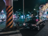 A black taxi is parked at a busy city intersection at night. Neon lights from surrounding buildings and advertisements illuminate the street. A large pillar with black and yellow stripes stands near the taxi. Several pedestrians are visible in the background, adding to the urban atmosphere.