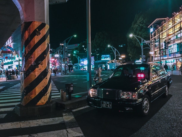 A black taxi is parked at a busy city intersection at night. Neon lights from surrounding buildings and advertisements illuminate the street. A large pillar with black and yellow stripes stands near the taxi. Several pedestrians are visible in the background, adding to the urban atmosphere.
