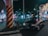 A black taxi is parked at a busy city intersection at night. Neon lights from surrounding buildings and advertisements illuminate the street. A large pillar with black and yellow stripes stands near the taxi. Several pedestrians are visible in the background, adding to the urban atmosphere.