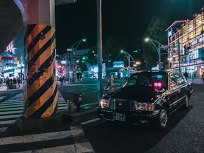 A black taxi is parked at a busy city intersection at night. Neon lights from surrounding buildings and advertisements illuminate the street. A large pillar with black and yellow stripes stands near the taxi. Several pedestrians are visible in the background, adding to the urban atmosphere.