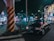 A black taxi is parked at a busy city intersection at night. Neon lights from surrounding buildings and advertisements illuminate the street. A large pillar with black and yellow stripes stands near the taxi. Several pedestrians are visible in the background, adding to the urban atmosphere.