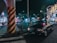 A black taxi is parked at a busy city intersection at night. Neon lights from surrounding buildings and advertisements illuminate the street. A large pillar with black and yellow stripes stands near the taxi. Several pedestrians are visible in the background, adding to the urban atmosphere.
