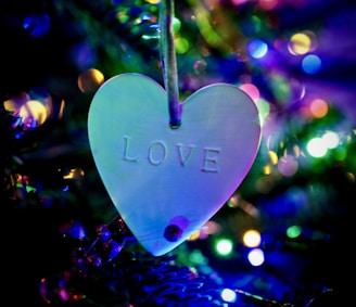A heart-shaped ornament with the word 'LOVE' embossed on it hangs on a string. The background features colorful, blurry lights, creating a festive and warm atmosphere.