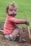 A young child with curly hair sits on the ground, covered in dirt, particularly on the face and hands. The child is wearing a pink long-sleeve shirt and a diaper, holding a wooden stick. They appear to be enjoying outdoor play, with a grassy background visible.