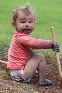 A young child with curly hair sits on the ground, covered in dirt, particularly on the face and hands. The child is wearing a pink long-sleeve shirt and a diaper, holding a wooden stick. They appear to be enjoying outdoor play, with a grassy background visible.