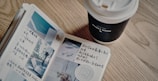 An open journal with handwritten notes and small photographs is placed on a wooden table. Next to it is a takeaway coffee cup with a lid, labeled with the words 'Honey Wood'.