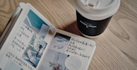 An open journal with handwritten notes and small photographs is placed on a wooden table. Next to it is a takeaway coffee cup with a lid, labeled with the words 'Honey Wood'.