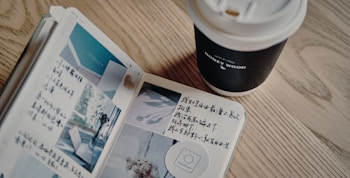An open journal with handwritten notes and small photographs is placed on a wooden table. Next to it is a takeaway coffee cup with a lid, labeled with the words 'Honey Wood'.