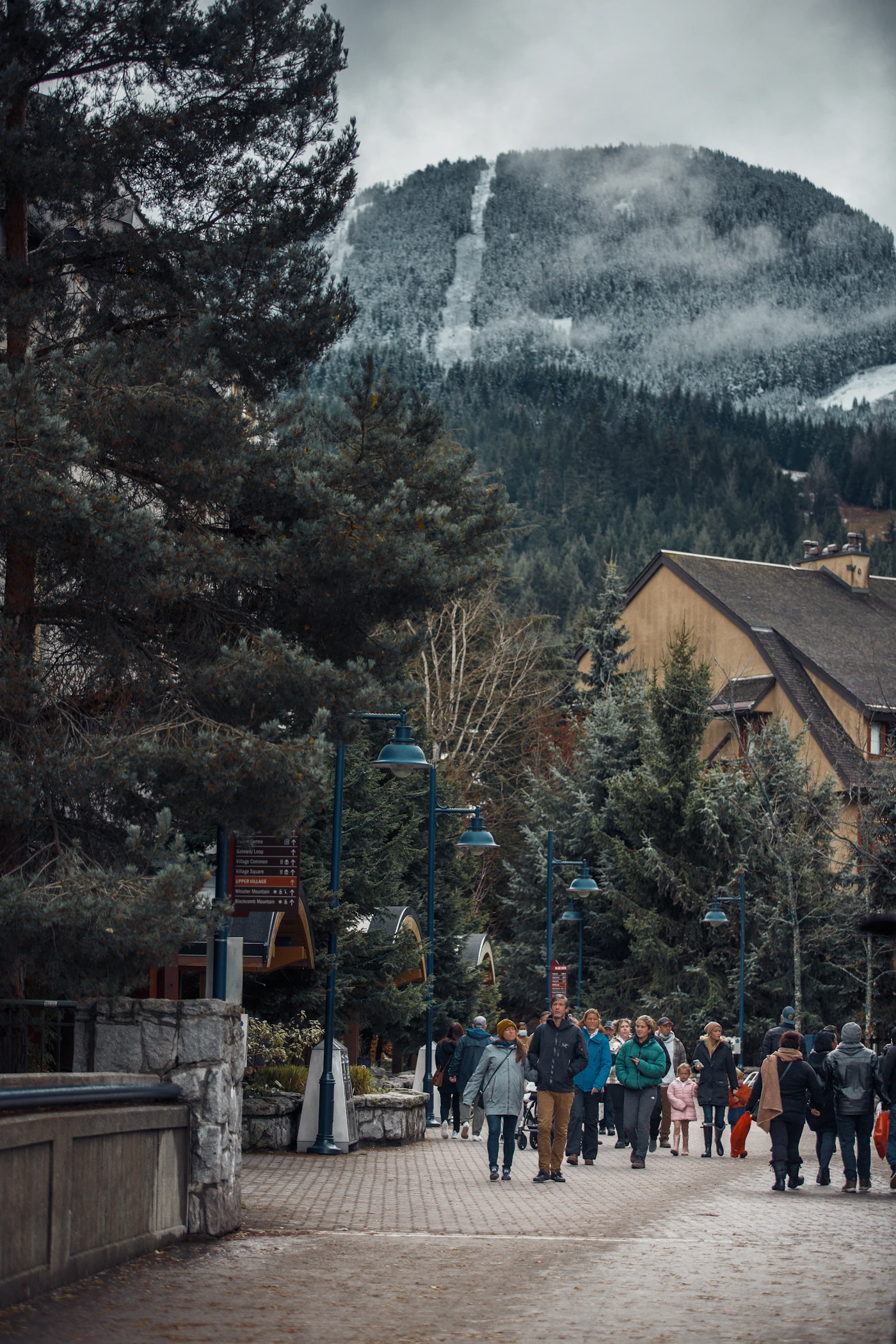 Whistler village pedestrian promenade in winter, people walking past timber-frame resort buildings with a snow-covered mountain rising directly behind