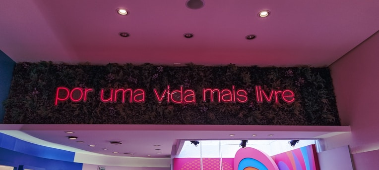 A neon sign displaying the phrase 'por uma vida mais livre' in pink lights is mounted on a wall covered with green foliage. Below the sign, a ceiling with recessed lighting features blue and pink accents.