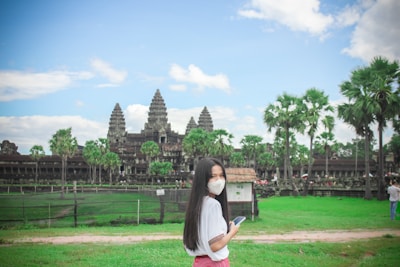 A person wearing a white mask and holding a smartphone stands on a green grassy area in front of a historical temple complex with tall spires. The sky is blue with some clouds, and there are several palm trees in the surroundings.