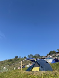 A sunny outdoor camping scene with multiple tents set up on a grassy hillside under a clear blue sky. The foreground features a prominent blue and yellow tent, while other tents and camping gear are visible in the background. Some trees and shrubbery dot the landscape, enhancing the natural setting.
