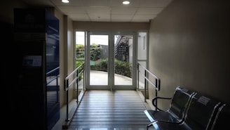A well-lit hallway with a glass door at the end, leading to an outdoor area with greenery. The space is equipped with metal railings and metallic chairs on the side. An ATM is visible on the left side, with the logo of a financial institution.