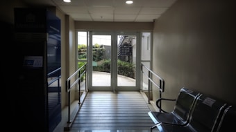 A well-lit hallway with a glass door at the end, leading to an outdoor area with greenery. The space is equipped with metal railings and metallic chairs on the side. An ATM is visible on the left side, with the logo of a financial institution.