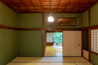 A traditional Japanese room featuring tatami mats on the floor and sliding shoji doors with wooden frames. The walls are painted in a muted green tone, and the ceiling has a wooden lattice design. A soft hanging light fixture provides illumination, while a view of a lush garden is visible through the open sliding door, adding a tranquil natural element to the space.