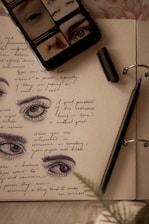 A journal page with sketches of eyes accompanied by handwritten notes in blue ink. A black pen lies on the open pages, with its cap nearby. A mobile phone displaying images of eyes rests on the journal.