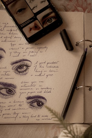 A journal page with sketches of eyes accompanied by handwritten notes in blue ink. A black pen lies on the open pages, with its cap nearby. A mobile phone displaying images of eyes rests on the journal.