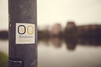 A sticker with the text 'Business Nomad' is affixed to a weathered metallic pole. In the background, an out-of-focus view of a riverfront cityscape with buildings and their reflections in the water.