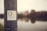A sticker with the text 'Business Nomad' is affixed to a weathered metallic pole. In the background, an out-of-focus view of a riverfront cityscape with buildings and their reflections in the water.