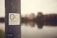 A sticker with the text 'Business Nomad' is affixed to a weathered metallic pole. In the background, an out-of-focus view of a riverfront cityscape with buildings and their reflections in the water.