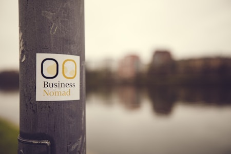 A sticker with the text 'Business Nomad' is affixed to a weathered metallic pole. In the background, an out-of-focus view of a riverfront cityscape with buildings and their reflections in the water.