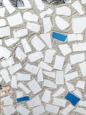 A mosaic pattern featuring irregularly shaped white tiles with scattered blue tiles set in a cement background. The texture gives a rough, handcrafted appearance with natural wear visible.