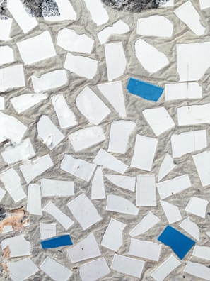 A mosaic pattern featuring irregularly shaped white tiles with scattered blue tiles set in a cement background. The texture gives a rough, handcrafted appearance with natural wear visible.