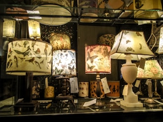 A collection of decorative table lamps displayed on a glass shelf, featuring shades adorned with intricate bird and butterfly patterns. The warm glow casts an inviting ambiance.