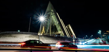 A striking modern church with an angular, triangular design is illuminated at night. Light trails from passing cars add dynamic elements to the scene, contrasting with the serene, snow-covered surroundings. A lit Christmas tree is visible inside the church through large windows, adding a festive touch.
