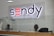 A reception desk with a glass barrier and wooden base is shown. Behind the desk, two individuals are seated, one of whom is wearing headphones. Above the desk, a large illuminated sign reads 'Sendy Partner Center.'