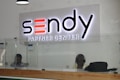 A reception desk with a glass barrier and wooden base is shown. Behind the desk, two individuals are seated, one of whom is wearing headphones. Above the desk, a large illuminated sign reads 'Sendy Partner Center.'