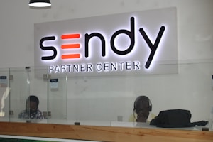 A reception desk with a glass barrier and wooden base is shown. Behind the desk, two individuals are seated, one of whom is wearing headphones. Above the desk, a large illuminated sign reads 'Sendy Partner Center.'