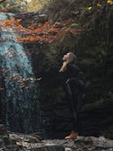 A woman stands confidently next to a waterfall surrounded by dense greenery. Autumn leaves provide a splash of orange against the lush green backdrop. The woman is dressed in dark clothing with sturdy brown boots, looking upwards, conveying a sense of freedom and connection with nature.