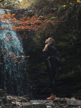 A woman stands confidently next to a waterfall surrounded by dense greenery. Autumn leaves provide a splash of orange against the lush green backdrop. The woman is dressed in dark clothing with sturdy brown boots, looking upwards, conveying a sense of freedom and connection with nature.