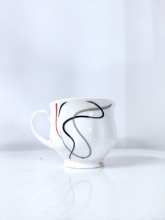A white ceramic cup with a curvy abstract design in black and red, placed on a glossy white surface. The background is a seamless continuation of the same white surface, creating a simple and minimalistic appearance.