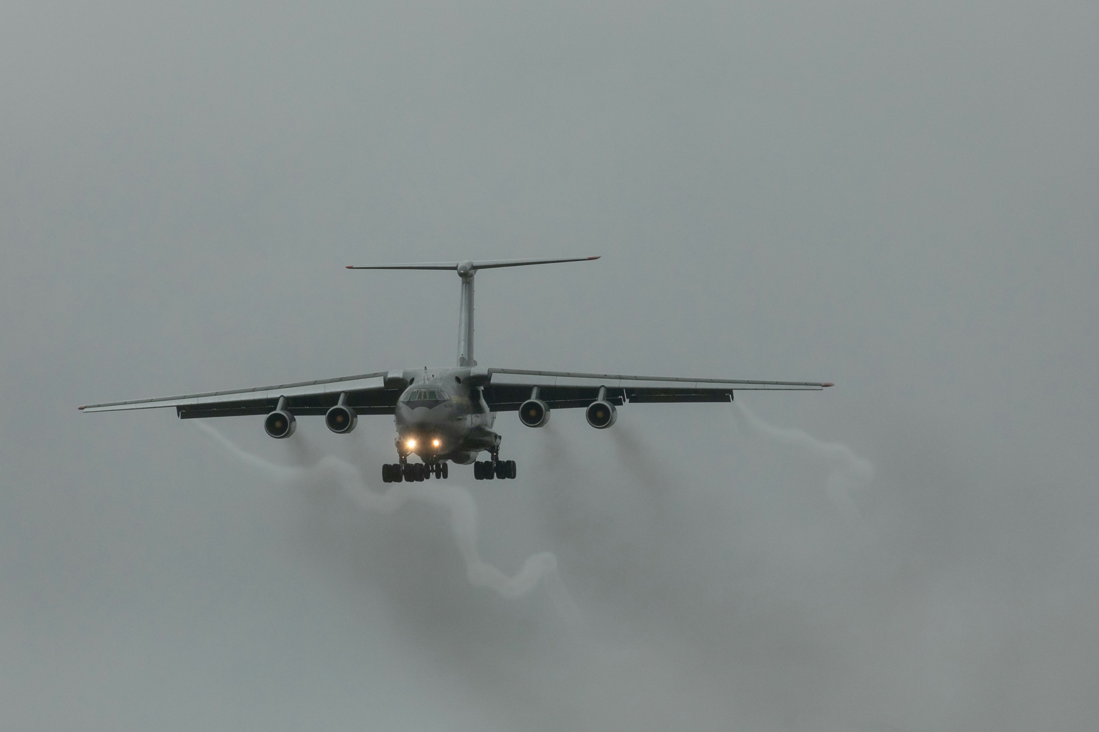 Ilyushin-76 approaching for landing with landing lights on, emitting smoke trails against a cloudy grey sky.