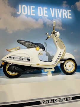 A stylish white Vespa scooter designed in collaboration with Christian Dior is displayed against a backdrop of blue sky and clouds. Golden accents and intricate patterns enhance its luxurious appearance. The slogan 'Joie de Vivre' is prominently displayed above.