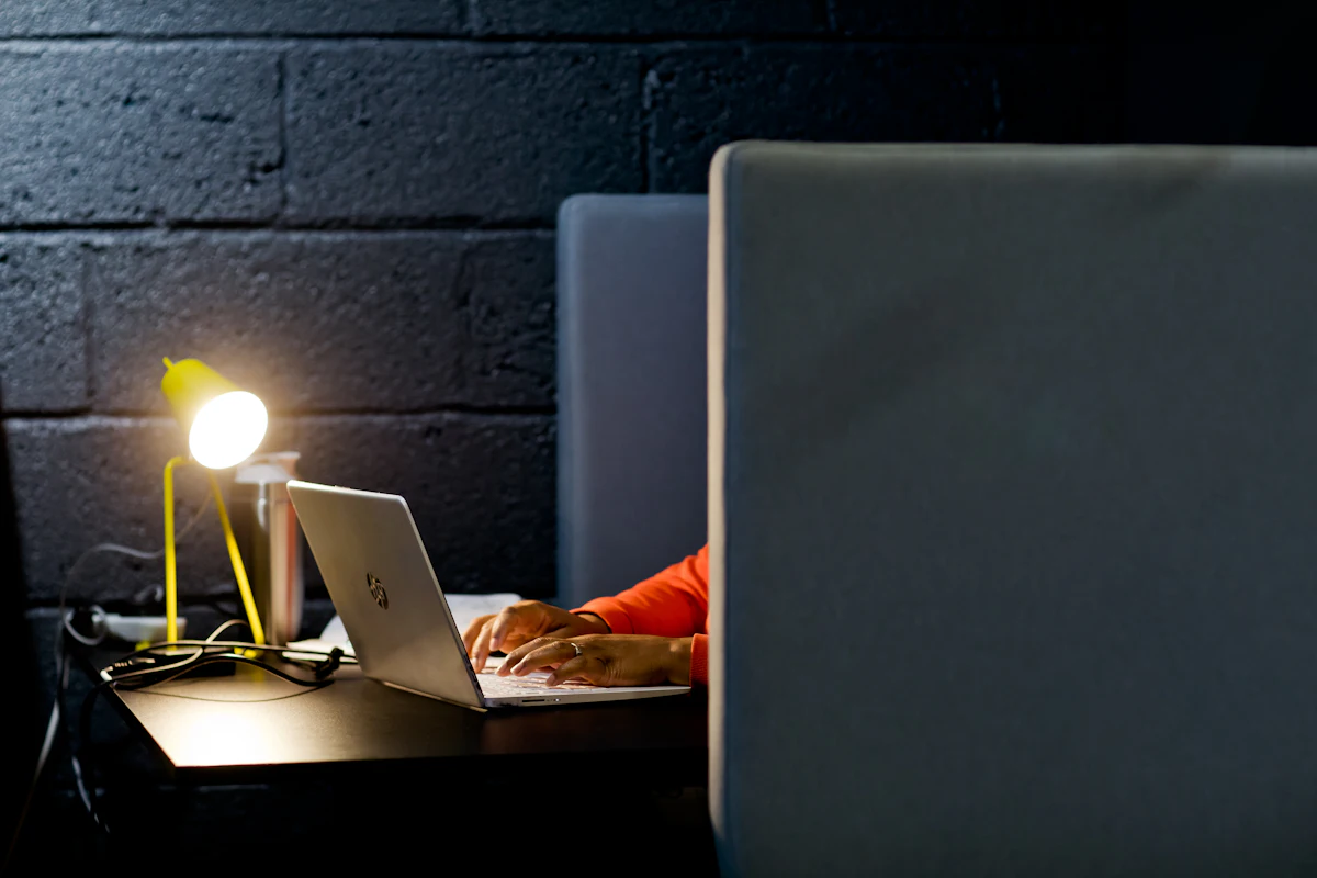 Seseorang mengetik di laptop pada malam hari di bawah cahaya lampu meja kuning yang hangat, dokumen kerja di sebelah tangan