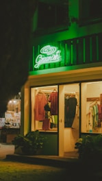 A warmly lit clothing store displays an array of garments on hangers, with a prominent green neon sign reading 'Greenich' above the entrance. The shop features various clothing items, and the exterior is decorated with potted plants.