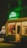 A warmly lit clothing store displays an array of garments on hangers, with a prominent green neon sign reading 'Greenich' above the entrance. The shop features various clothing items, and the exterior is decorated with potted plants.
