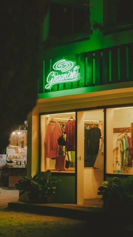 A warmly lit clothing store displays an array of garments on hangers, with a prominent green neon sign reading 'Greenich' above the entrance. The shop features various clothing items, and the exterior is decorated with potted plants.