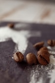 Coffee beans are scattered on a dark wooden surface with a dusting of white powder. The image has a shallow depth of field, highlighting the texture and details of the coffee beans in the foreground.
