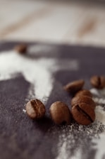 Coffee beans are scattered on a dark wooden surface with a dusting of white powder. The image has a shallow depth of field, highlighting the texture and details of the coffee beans in the foreground.