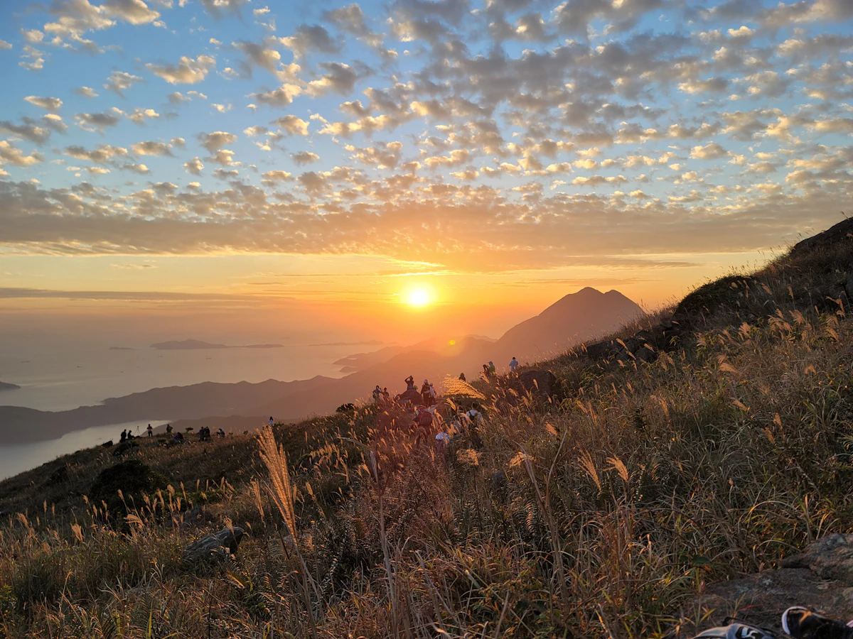 Hikers on the grassy slopes of Sunset Peak on Lantau Island at sunset, with a peak and the sea behind