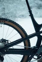 The image captures a close-up of the rear part of a bicycle. The bike's tire, chain, and frame are prominently visible. The bike is set against a snowy background with trees partially visible.