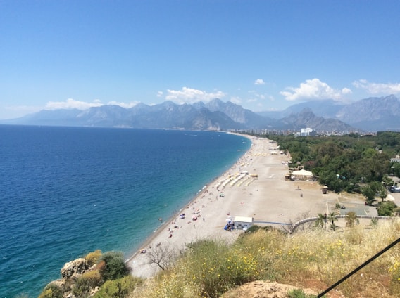 A vast coastline stretches alongside a clear blue sea with a pebble beach. The shoreline is lined with sun loungers and umbrellas. The background features a series of towering mountains beneath a partly cloudy sky. Lush greenery is present near the shore.