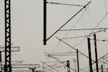 A complex network of silhouetted train electrical lines and poles with a light sky background, creating an intricate lattice of intersecting shapes against a pale backdrop.